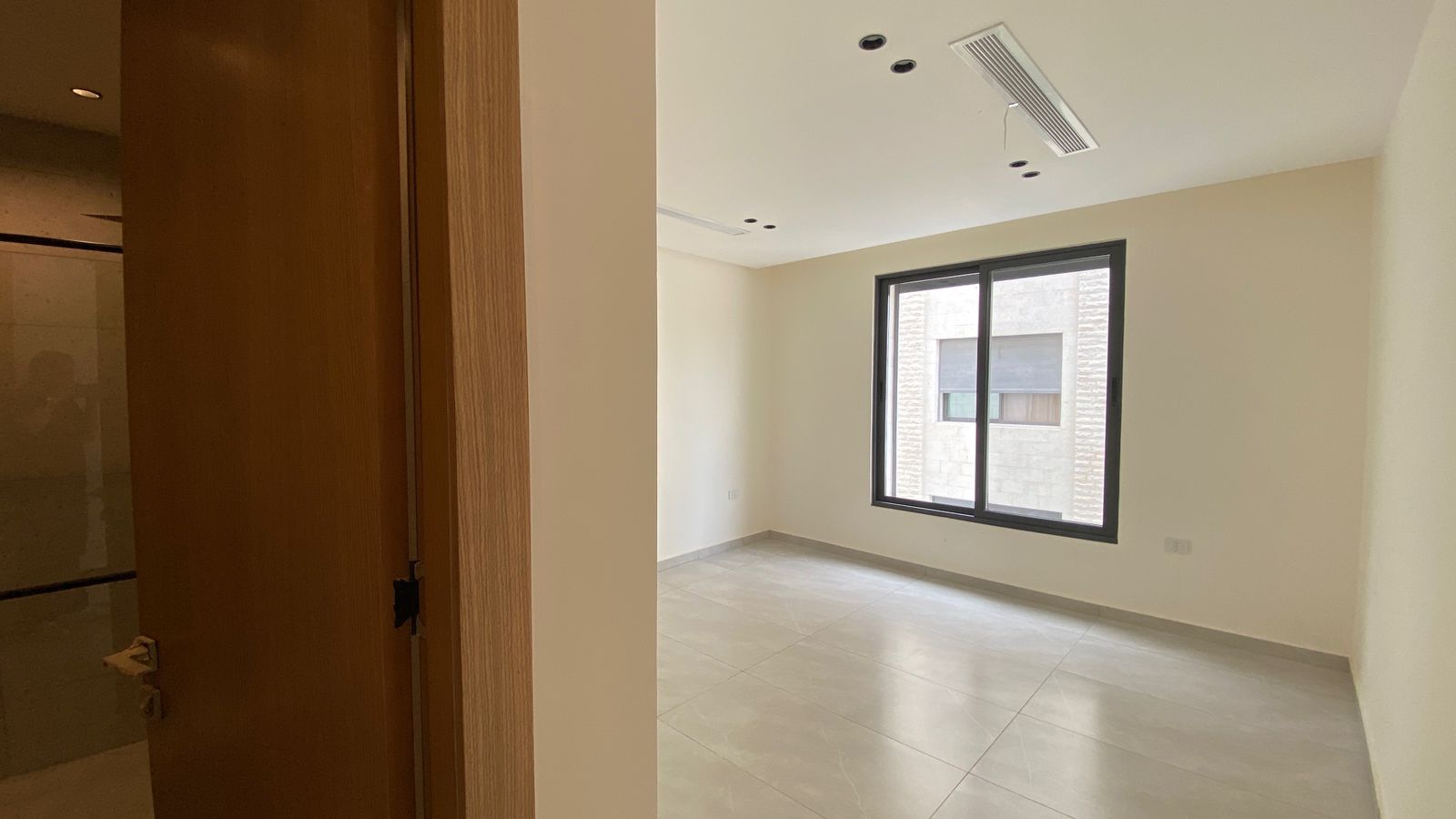 54a456c1-74fe-4c27-8cc2-c70bf14eef5c Empty modern room with a large window, beige walls, and light gray tiled floor; a wooden doorway is on the left.