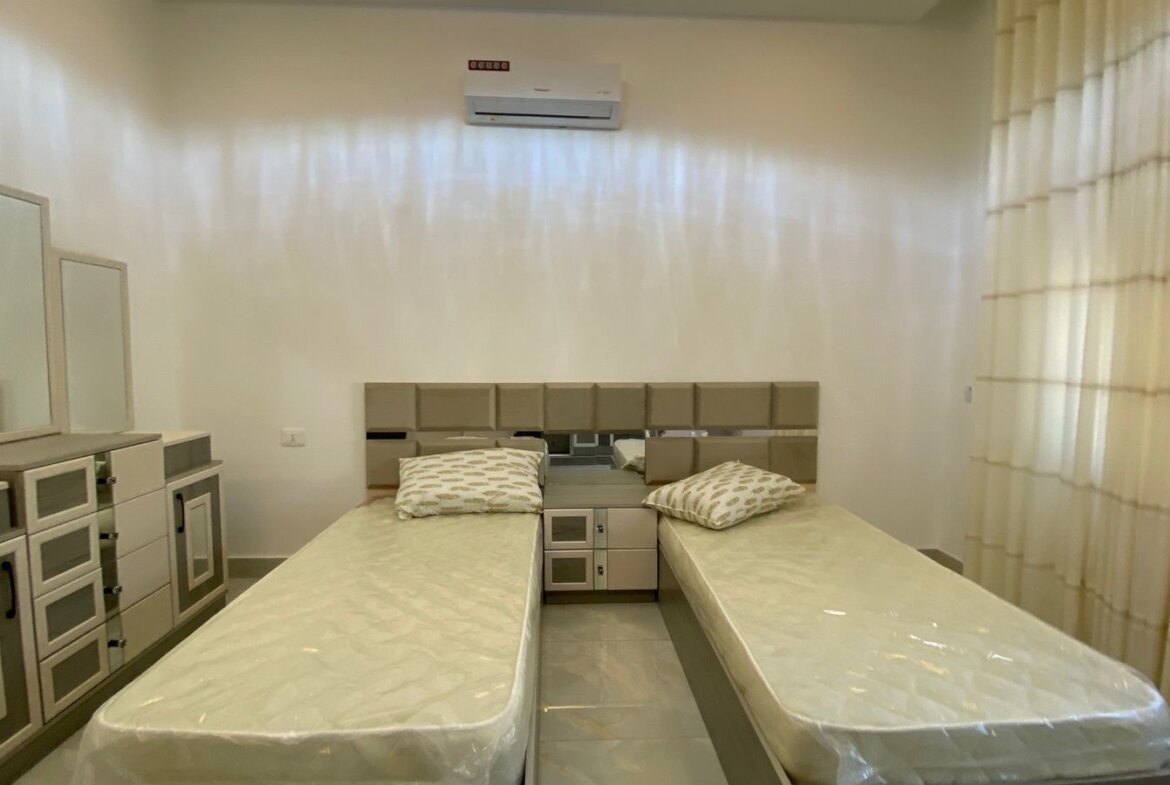 Twin beds in a minimalist hotel room with matching grey storage units and a shared nightstand between them, mirrors on the dresser to the left, and a wall-mounted air conditioner above.
