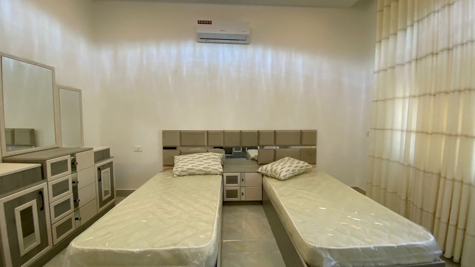 Twin beds in a minimalist hotel room with matching grey storage units and a shared nightstand between them, mirrors on the dresser to the left, and a wall-mounted air conditioner above.