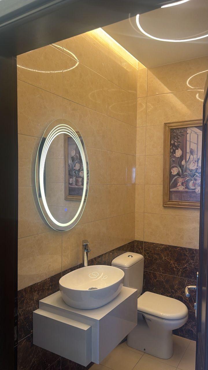 55a36d07-28bc-4cb4-bb08-18877bf6bfdf Modern bathroom with an oval LED-lit mirror, white vessel sink on a floating vanity, and a toilet against beige tile walls.