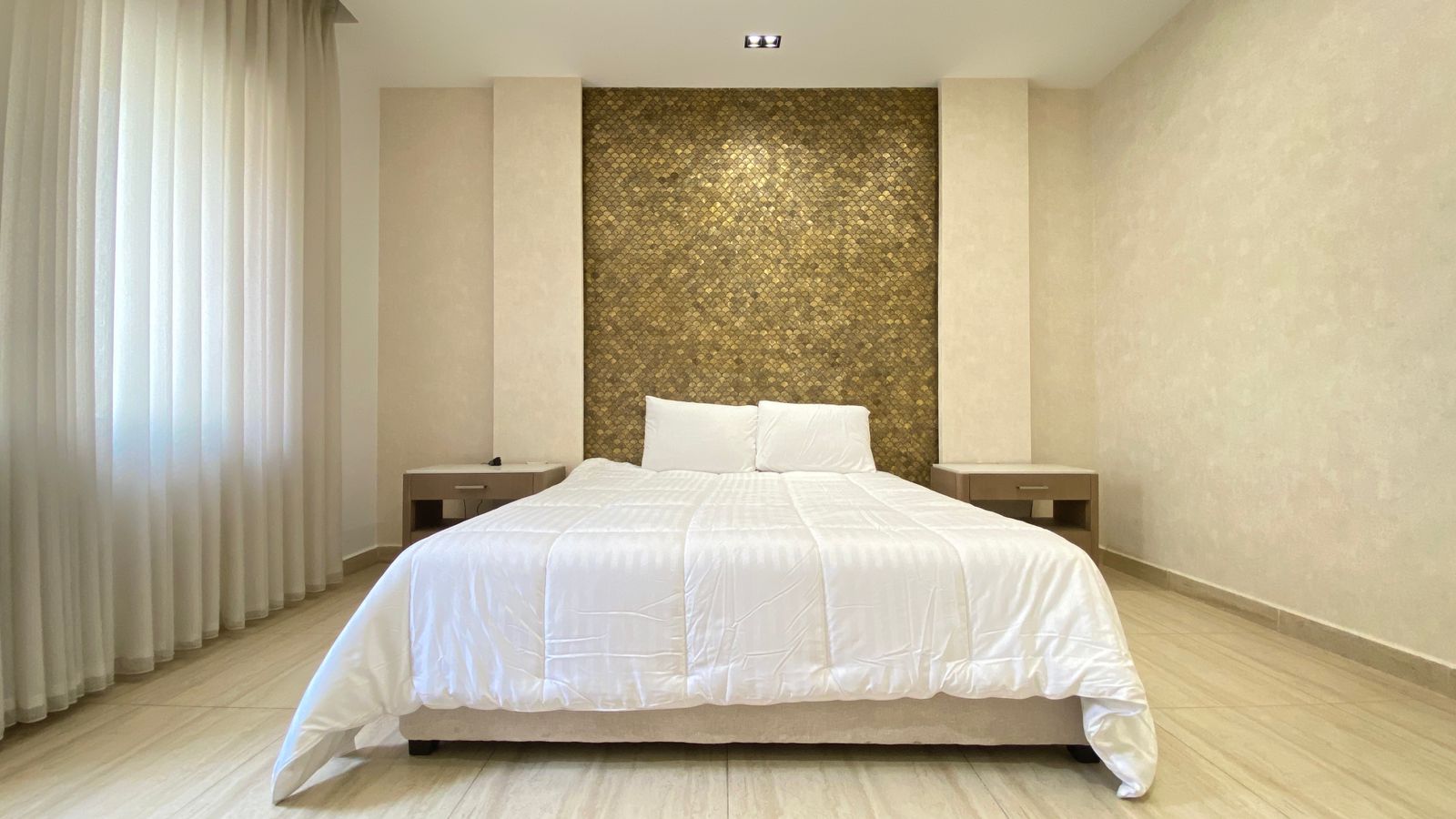 5621a031-4f4b-494a-9556-e6853618e952 Hotel bedroom with a white duvet, two pillows, and a mosaic brown headboard framed by beige walls and nightstands on each side.