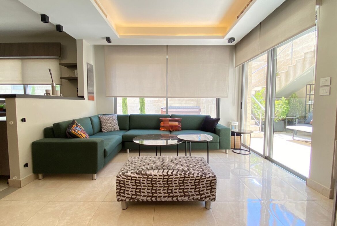 Open-concept living room with green sectional sofa, two round marble coffee tables, and a geometric ottoman; large windows with beige blinds