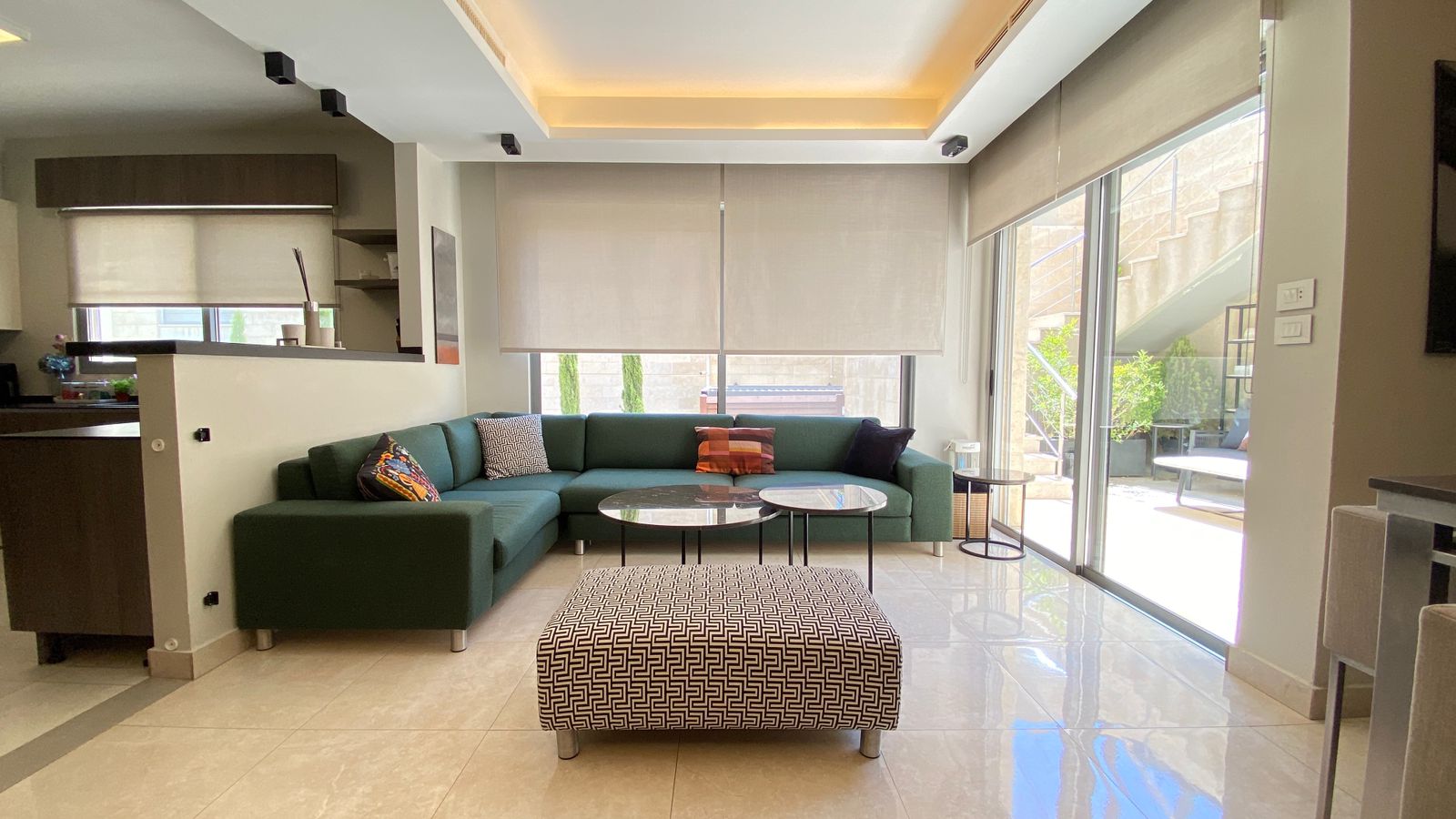 564c2060-904f-4dda-b0e9-158ed6b4c18b Open-concept living room with green sectional sofa, two round marble coffee tables, and a geometric ottoman; large windows with beige blinds