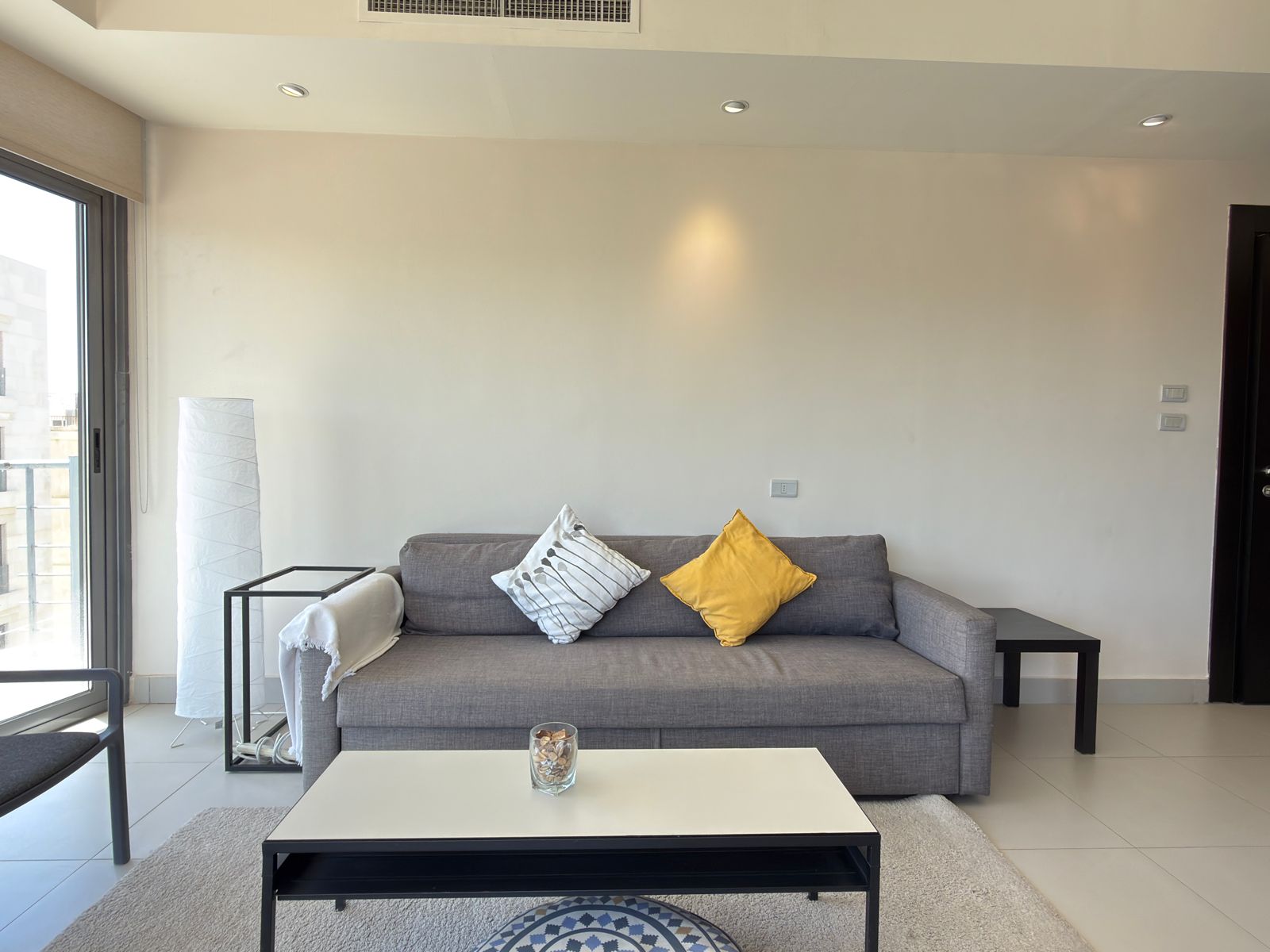56a27e27-d1df-44f8-9b23-4e960df1f315 Contemporary living room with a gray sofa, white walls, and two decorative cushions (yellow and striped) facing a white coffee table and black side tables on a light rug.