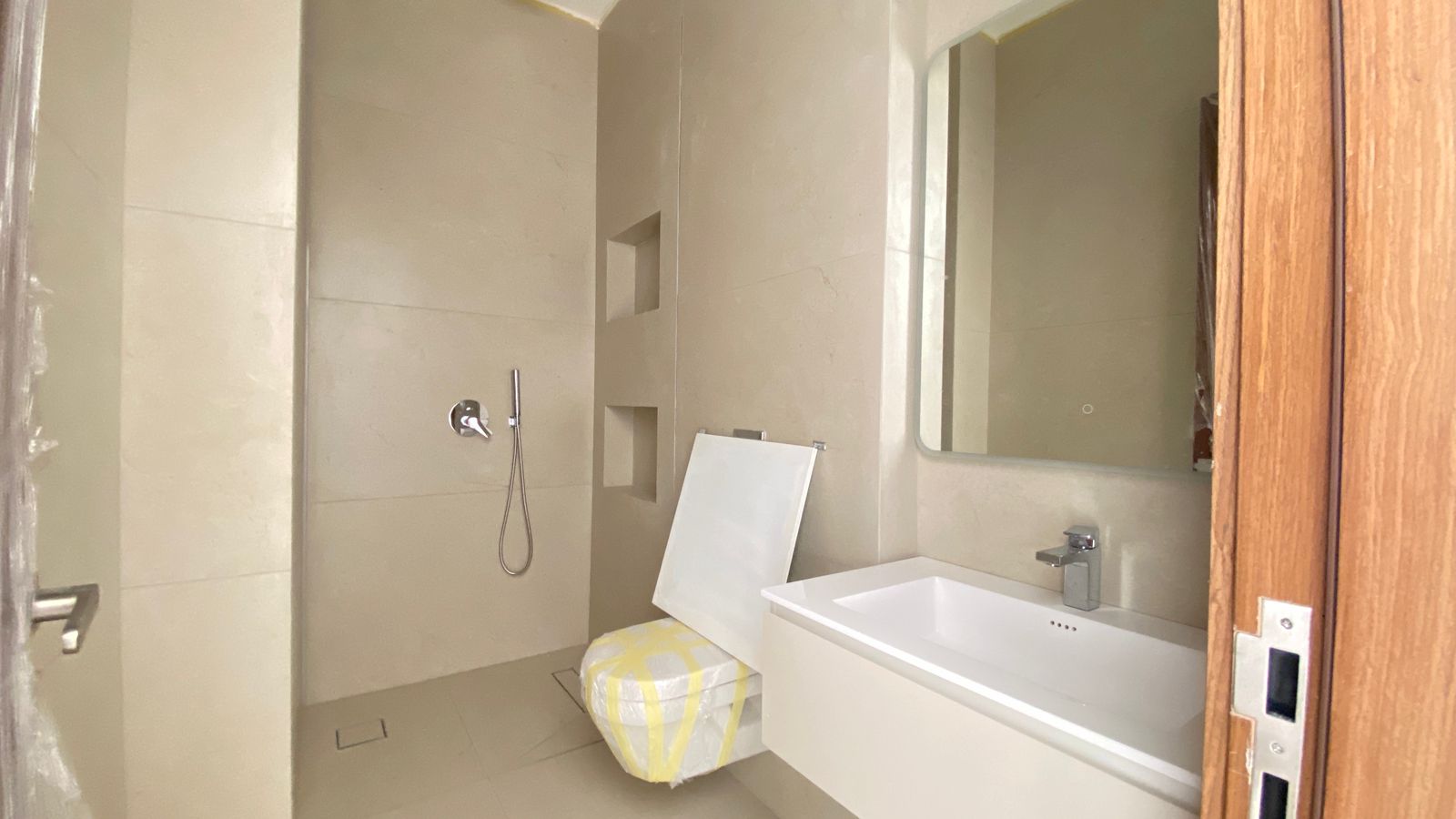 56ba2431-5db4-43c4-8b1f-6fe8742d3309 Beige-tiled bathroom under construction with wall niches, a white vanity with sink and large mirror, and a panel leaning on a wrapped stool near the shower area.