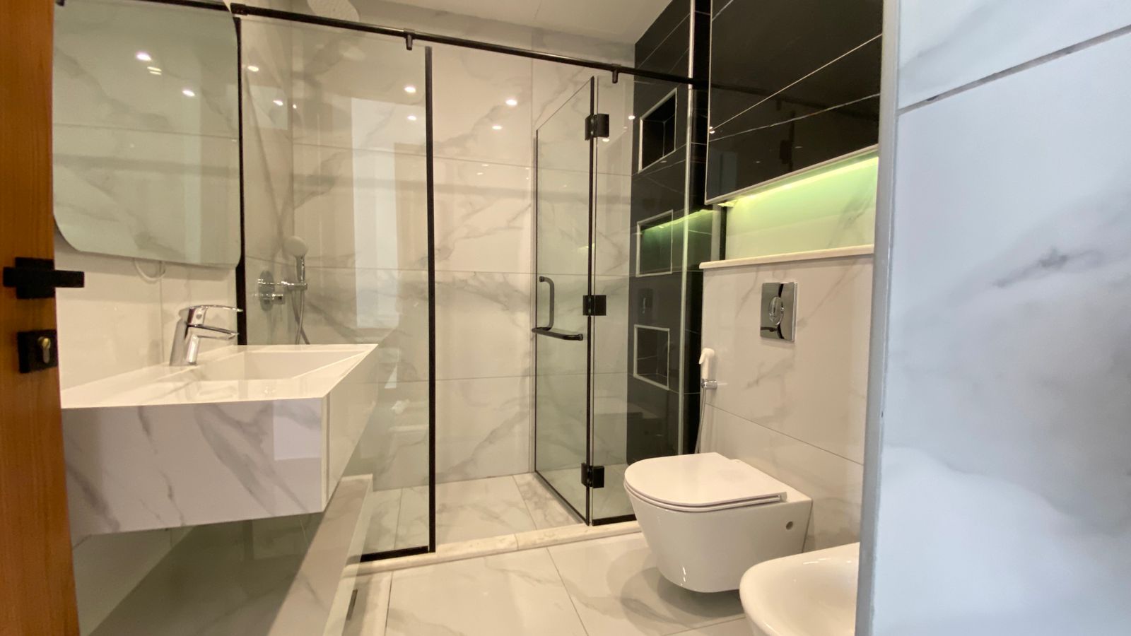 57043988-a4f7-4824-9771-9795eeffa492 Modern bathroom with a glass shower enclosure, wall-mounted white marble sink, and a wall-hung toilet; sleek, contemporary design.