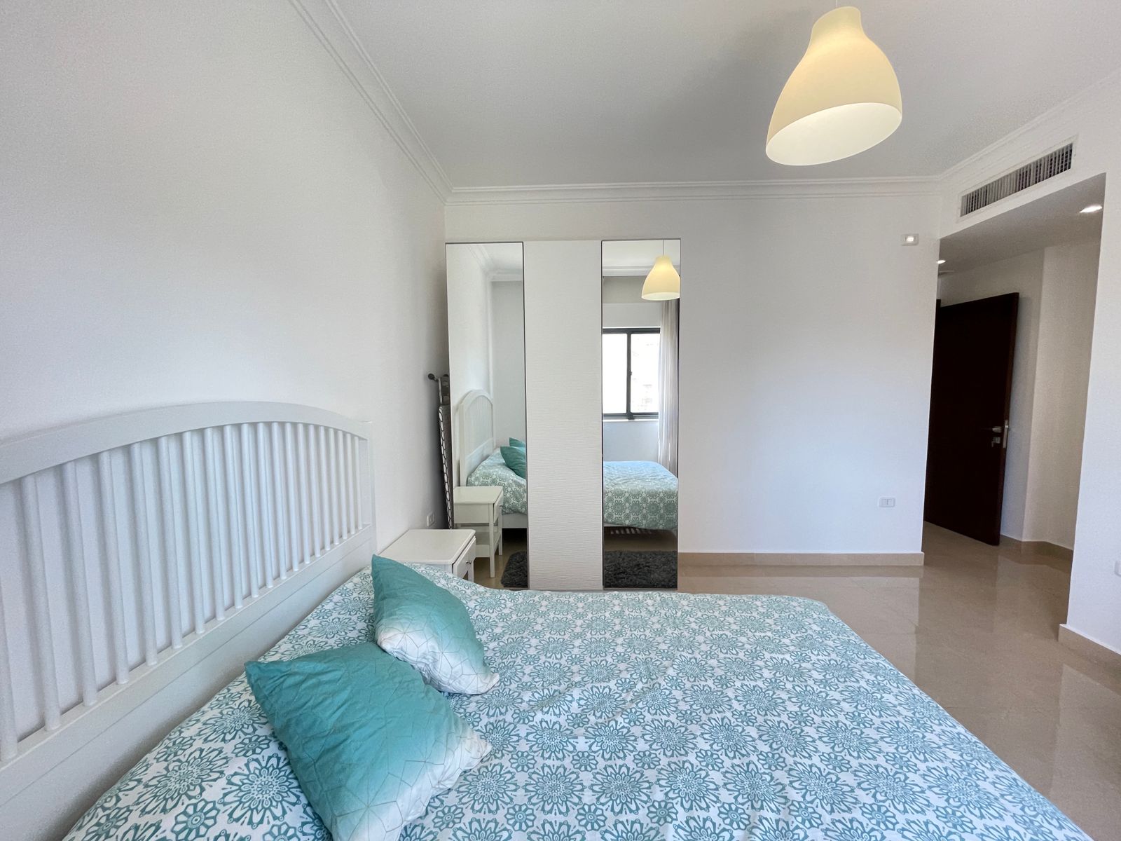 57927adb-974d-469f-b881-34f269cd84c9 Bright bedroom with white furniture, turquoise patterned bedding, and a mirrored wardrobe reflecting a window.