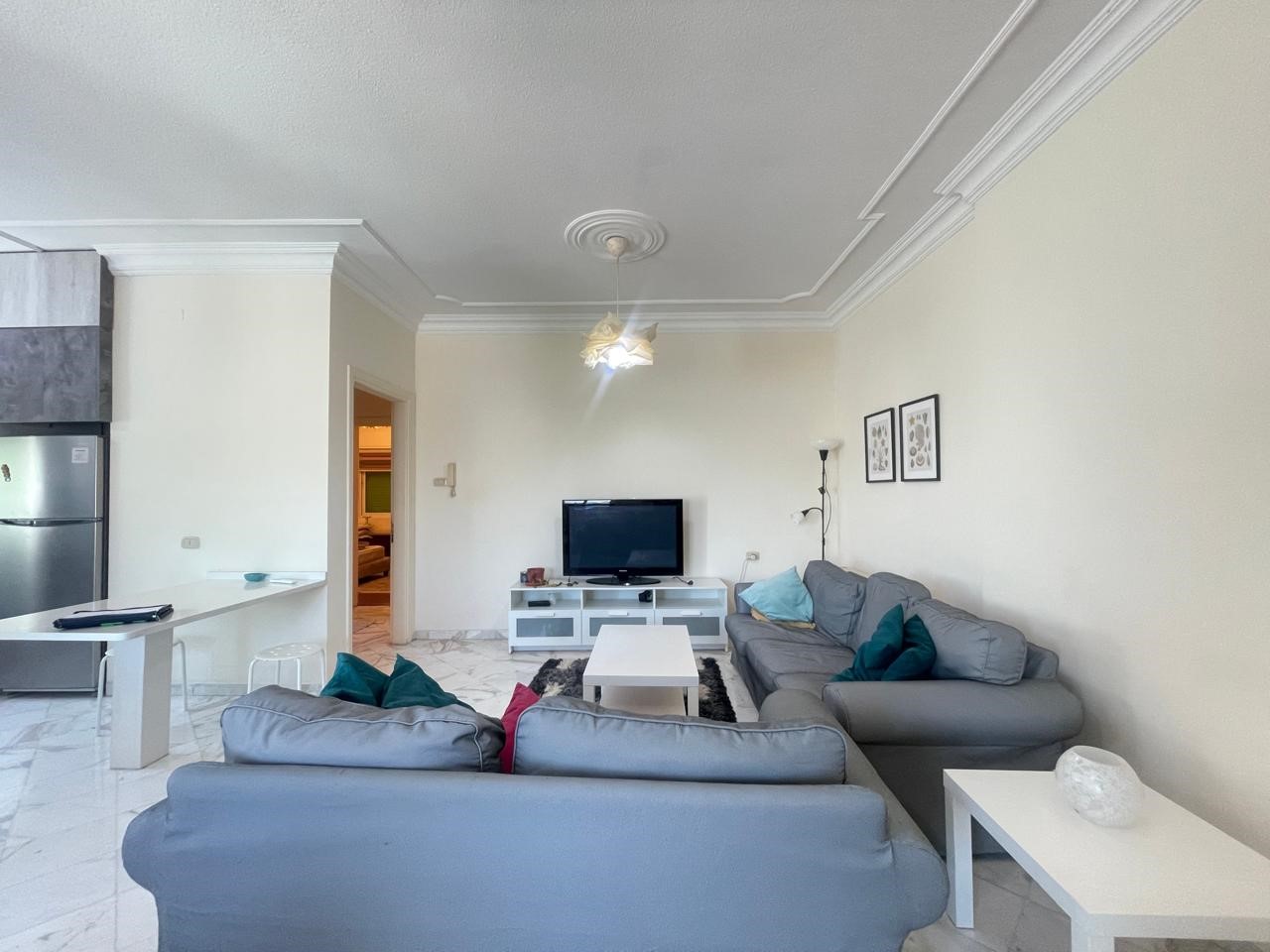 585df7b6-a66d-4e5b-add2-51d20dda96b0 Living room with two gray sectionals, white coffee table, and a TV stand against a pale wall; kitchen area visible to the left with a fridge and island.