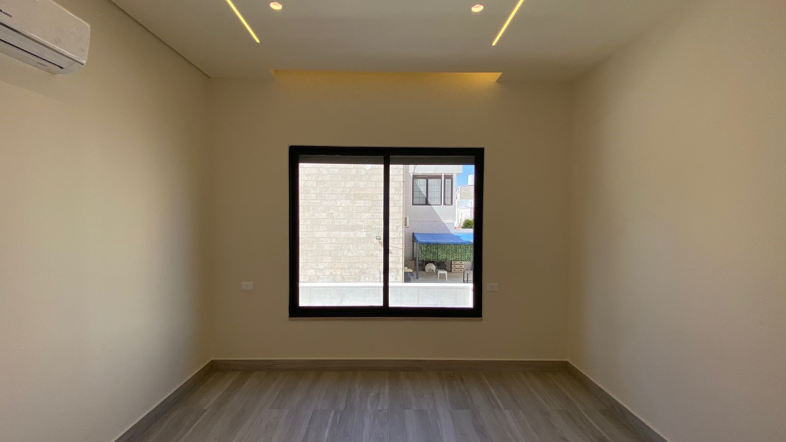 588e08ca-911e-4860-a82b-0de33ca34f55 Empty, unfurnished room with beige walls, wood-like floor, and a black-framed window center stage; wall-mounted AC on the left.