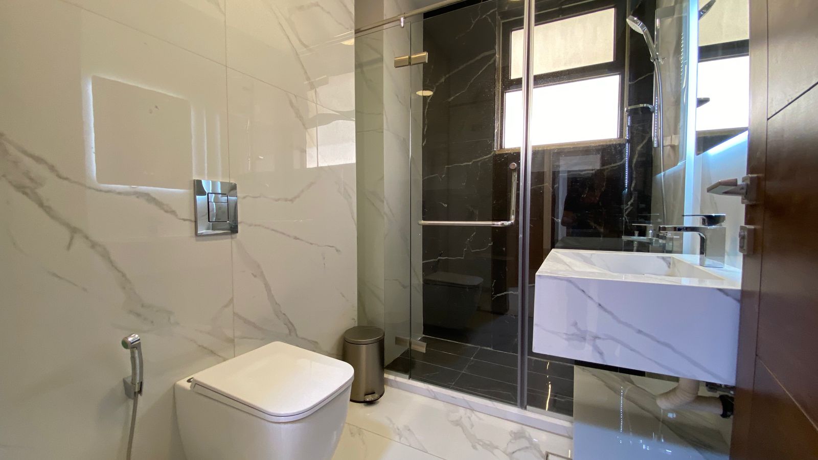 58f5a0c0-51cb-4385-bfc7-de9c8124cb4e Modern bathroom with white marble walls, glass shower enclosure, wall-mounted toilet, handheld bidet spray, and marble sink with chrome fixtures.