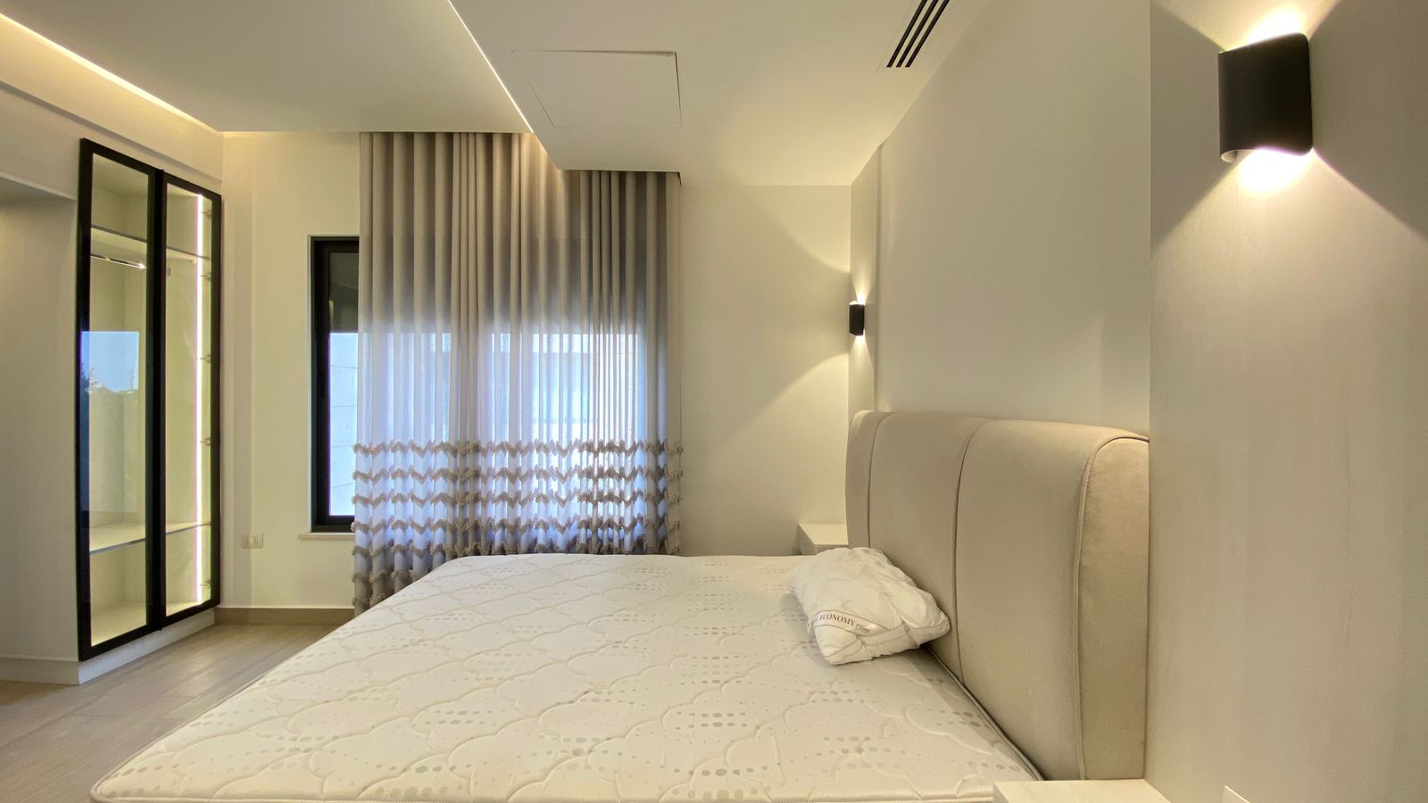 58f9e029-7f72-4ce5-a788-5025710dcc5d Modern minimalist bedroom with a beige upholstered bed, two white nightstands, wall sconces, and a glass-front wardrobe.