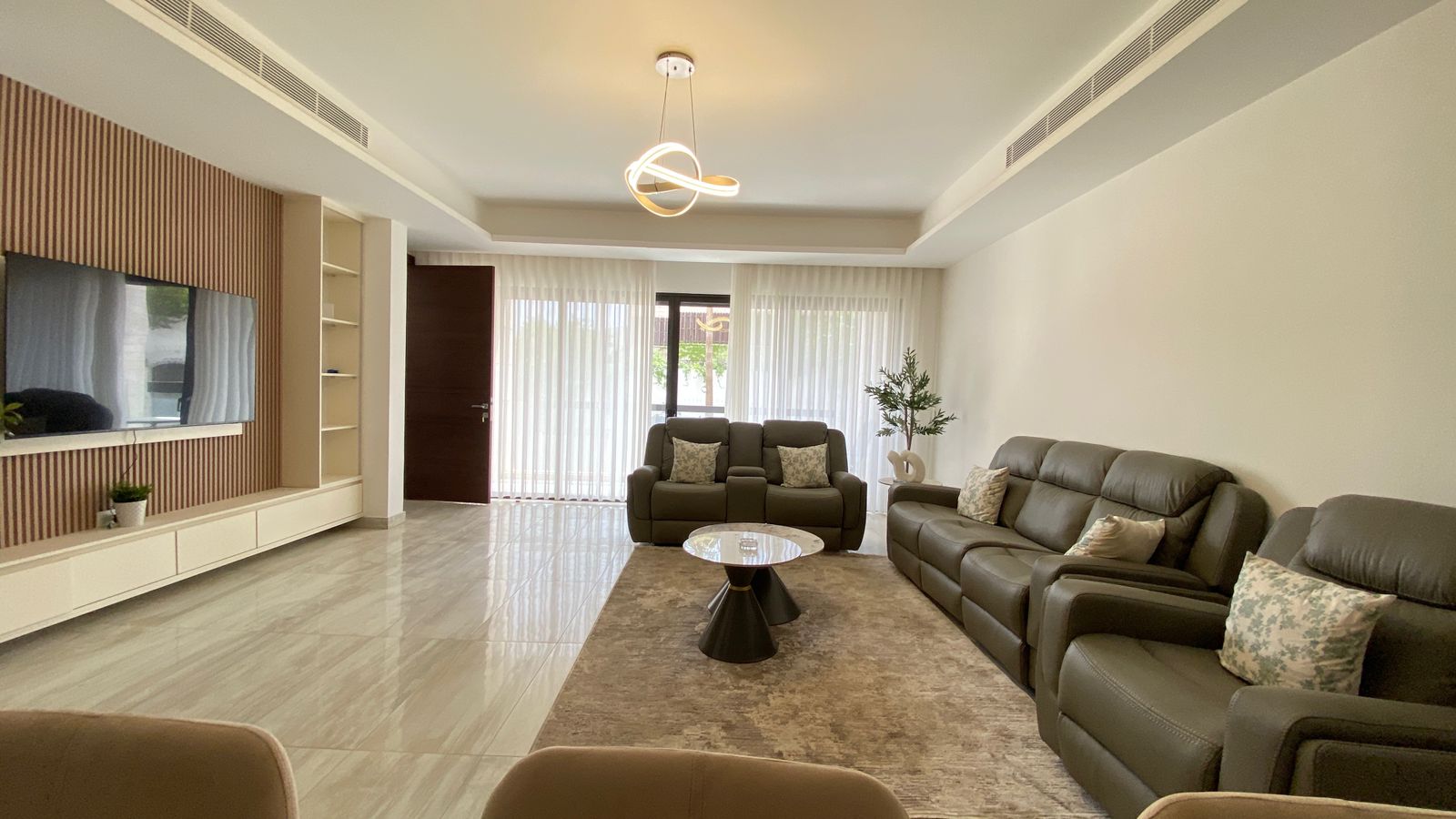 59c9263c-09ca-4c2b-94ac-3f4996b261f7 Contemporary living room with gray leather sofa set, glass-top coffee table on a beige rug, and a wall-mounted TV on a slatted wood wall used as a focal feature