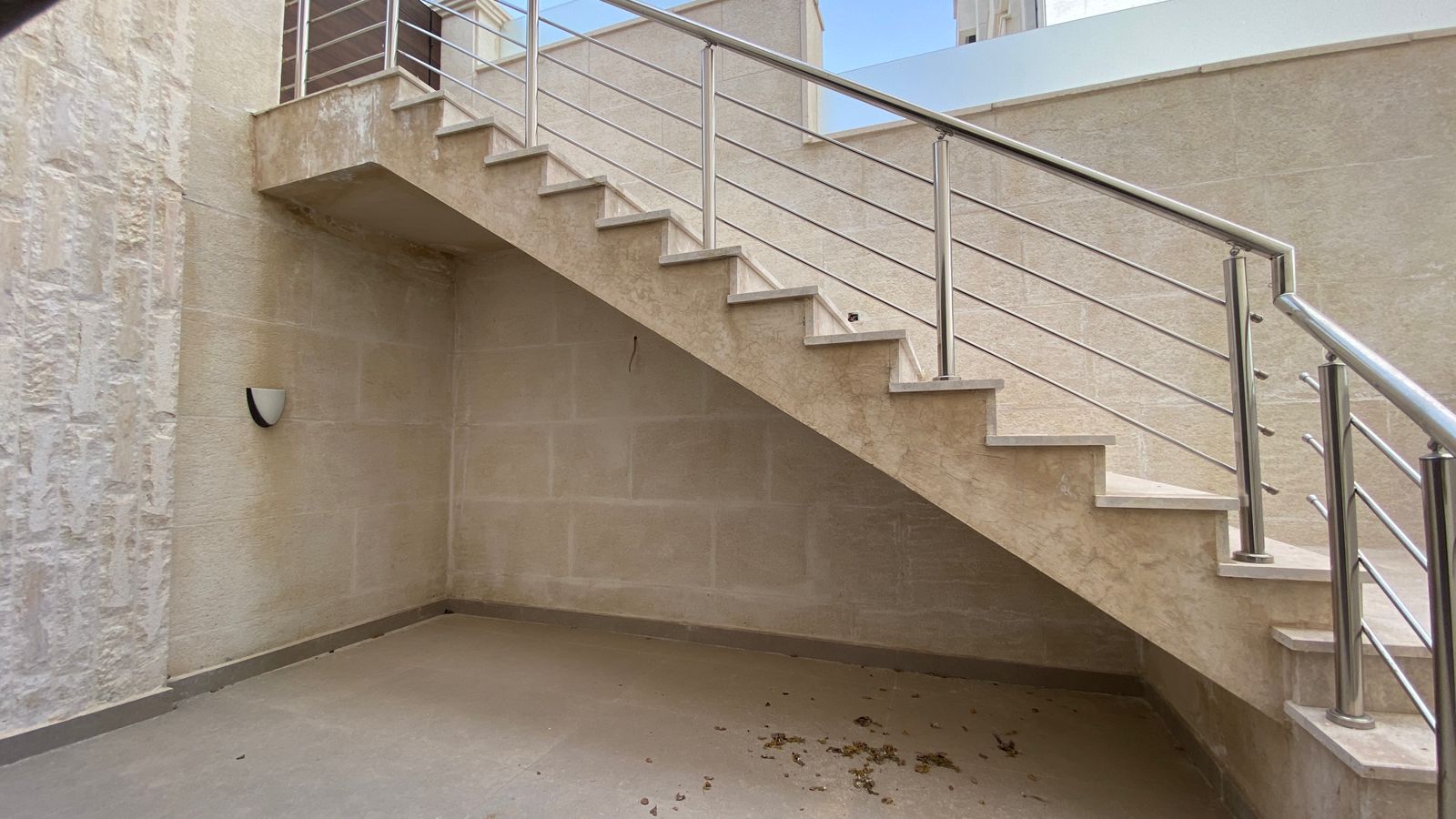 5a406b49-d8d9-47ea-a8fc-3340be8f6da3 Beige stone exterior staircase with stainless steel handrails along a wall; an open, shaded ground level beneath the steps remains clear of debris.