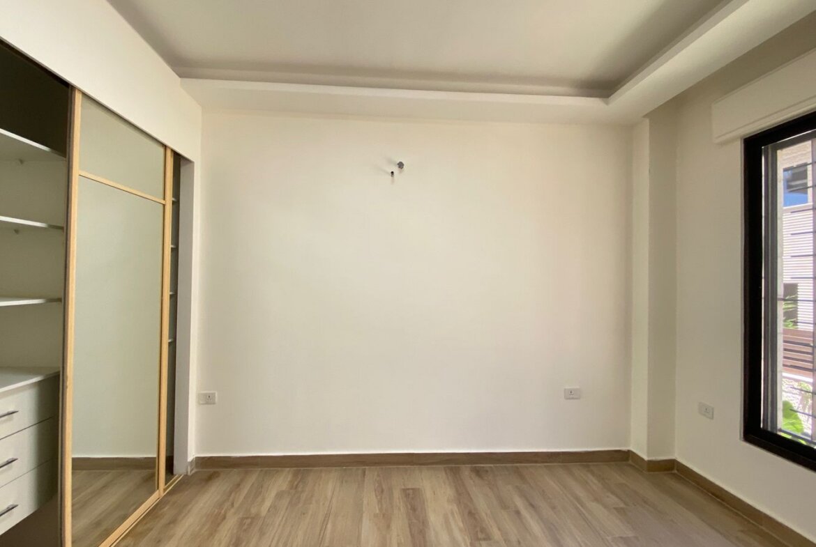 Empty minimalist room with light wood flooring, a built-in wardrobe with sliding mirror doors on the left, and a large window on the right.