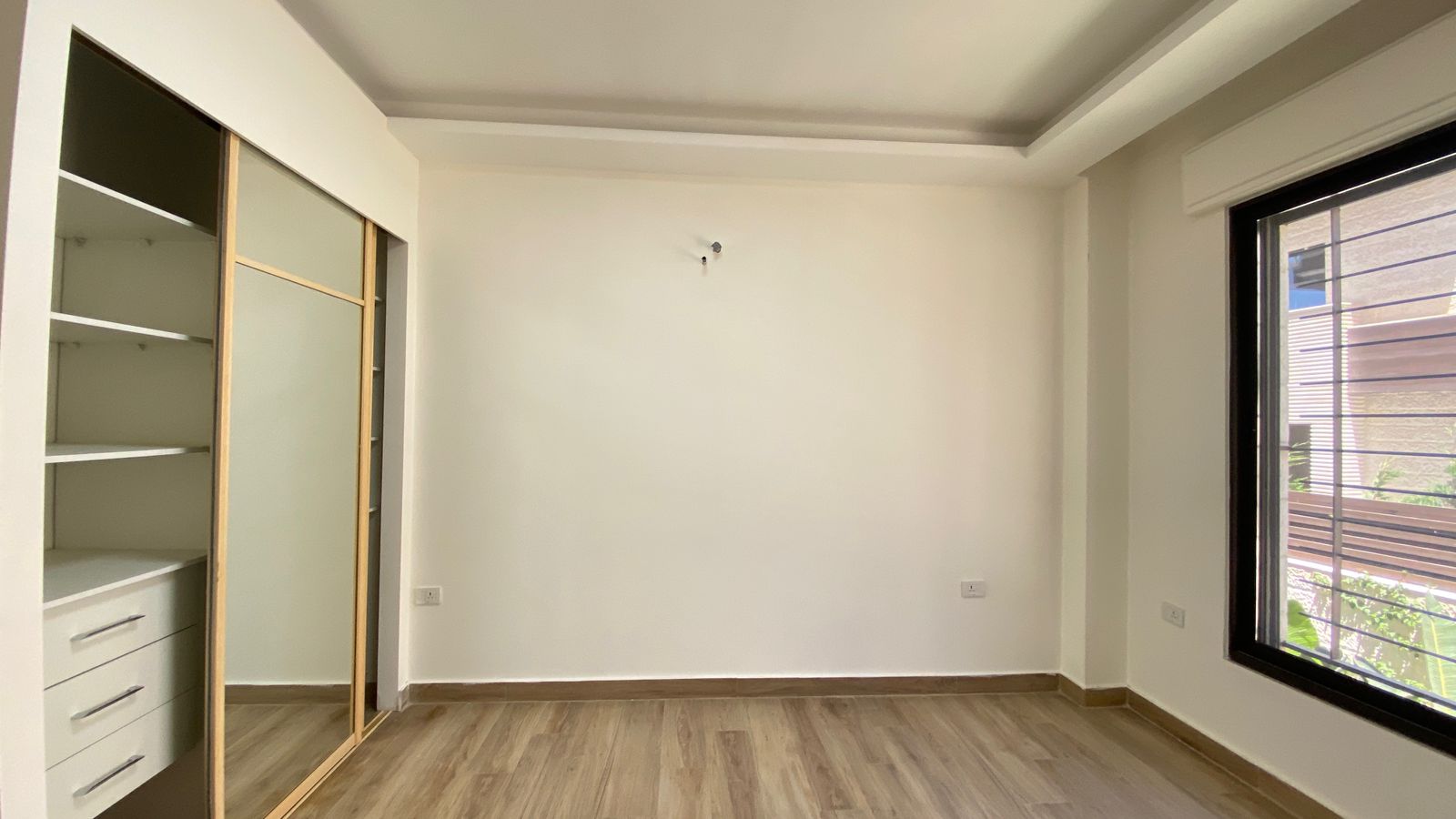 5a4ccbbd-aeed-4b00-a540-c2a84df8f7e1 Empty minimalist room with light wood flooring, a built-in wardrobe with sliding mirror doors on the left, and a large window on the right.