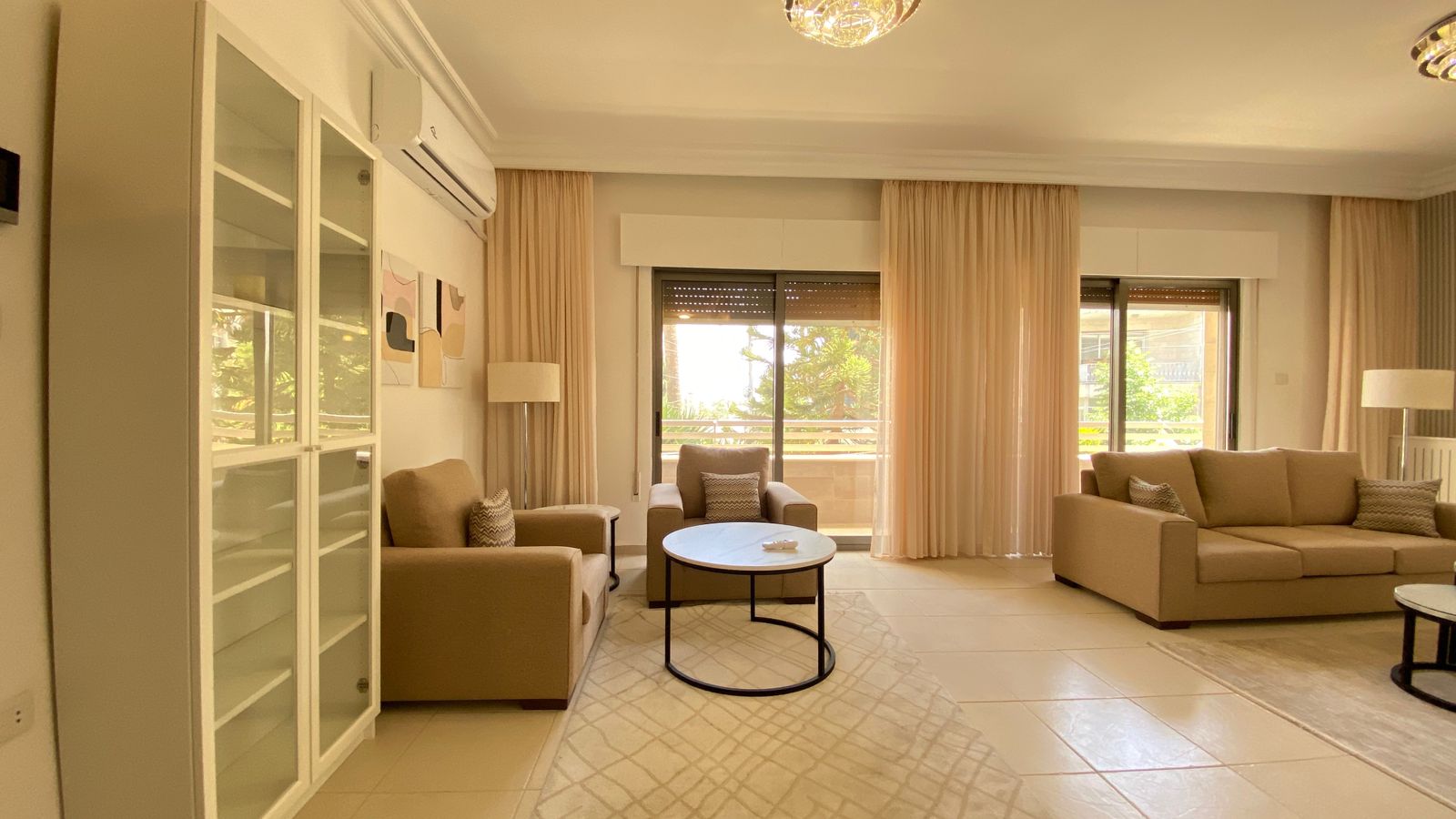 5acc26b5-841d-4a99-b264-d81dbb59797a Bright living room with beige sofas, a round coffee table, and a white cabinet along the left wall under an air conditioner, bathed in natural light from large windows.