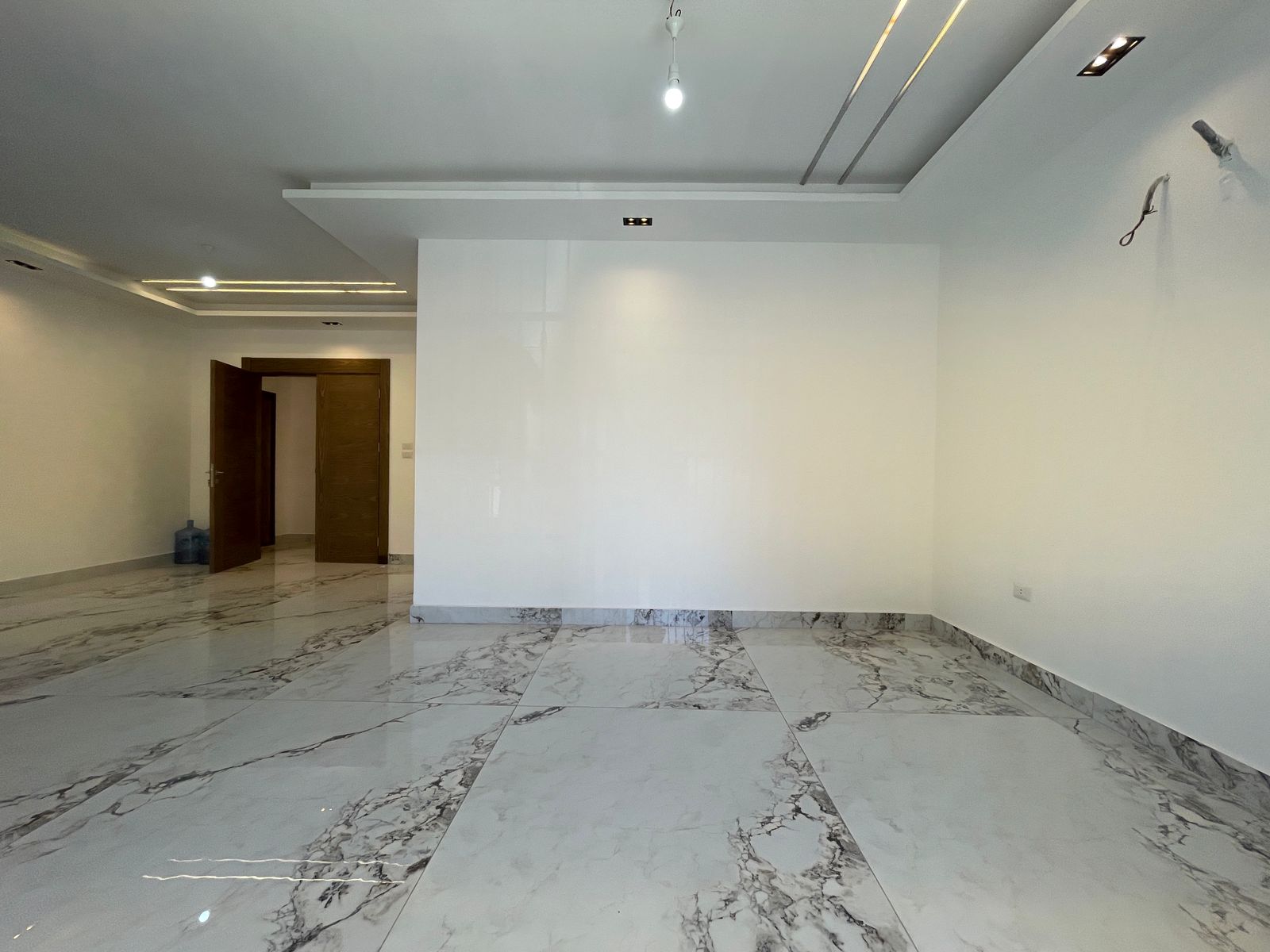 5b574886-d776-43a8-b32c-8ba60b75354f Empty modern room with white walls and marble floor; open wooden door leads to a hallway, with ceiling lights visible in the ceiling ridge.