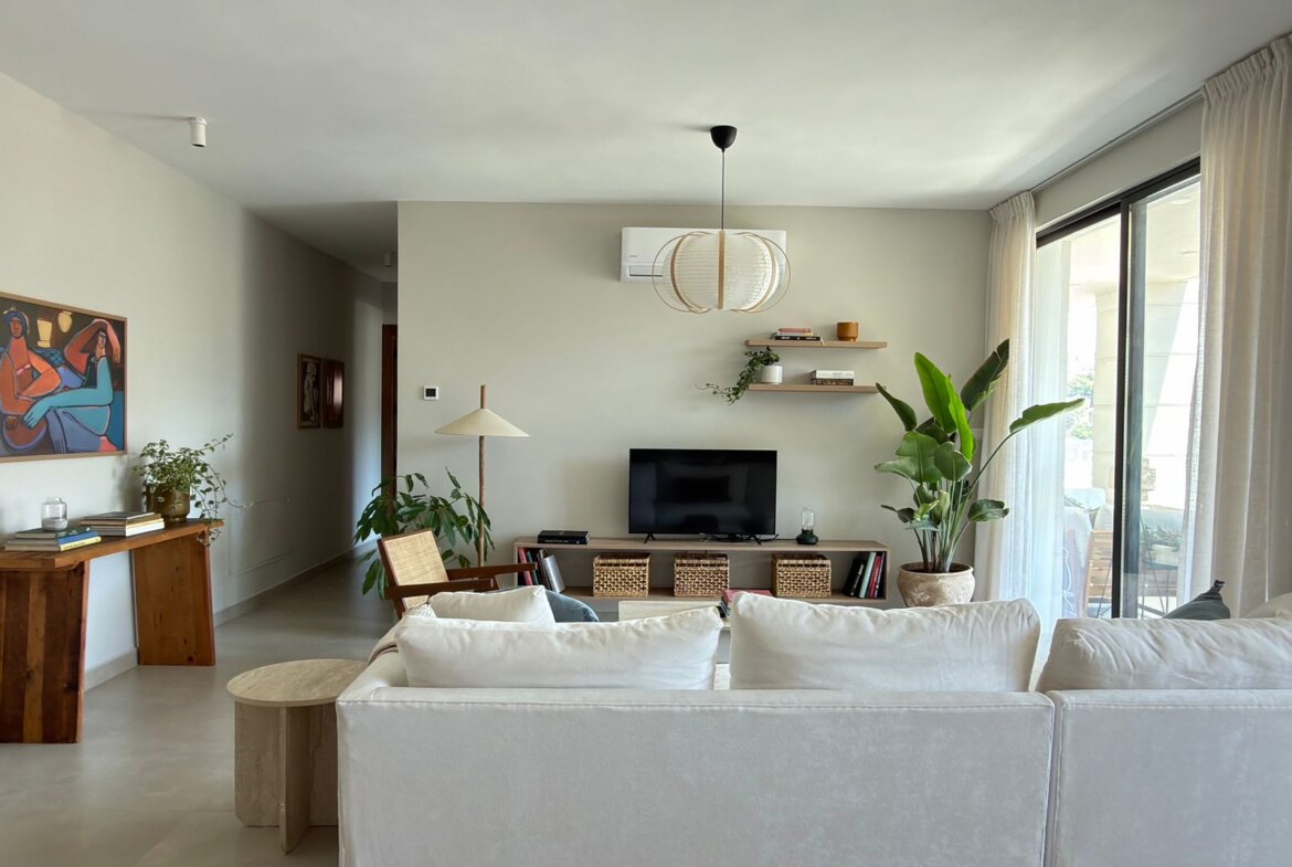 Bright living room with white sofa facing a TV, large sliding door with sheer curtains, and a few plants nearby.