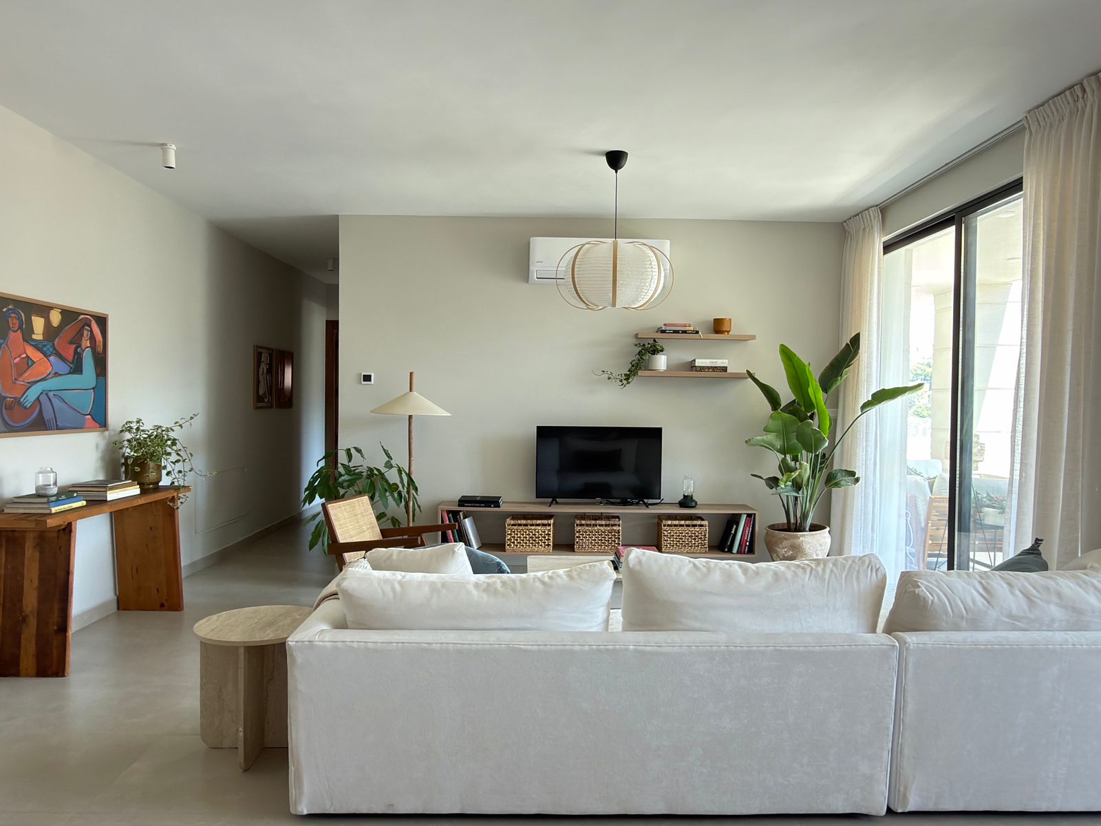 5c09a751-6482-4128-a520-d22f625e48de Bright living room with white sofa facing a TV, large sliding door with sheer curtains, and a few plants nearby.