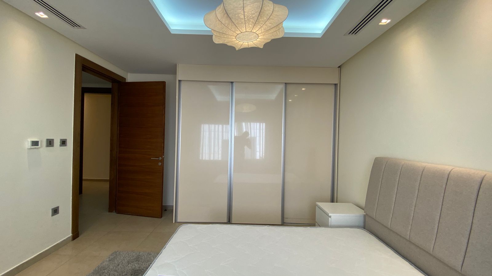 5c3166b4-3347-4e64-a165-a5b393994c2d Bedroom with a beige upholstered headboard, white nightstand, and large sliding-wardrobe doors; open wooden door to the hallway and a decorative ceiling light above.