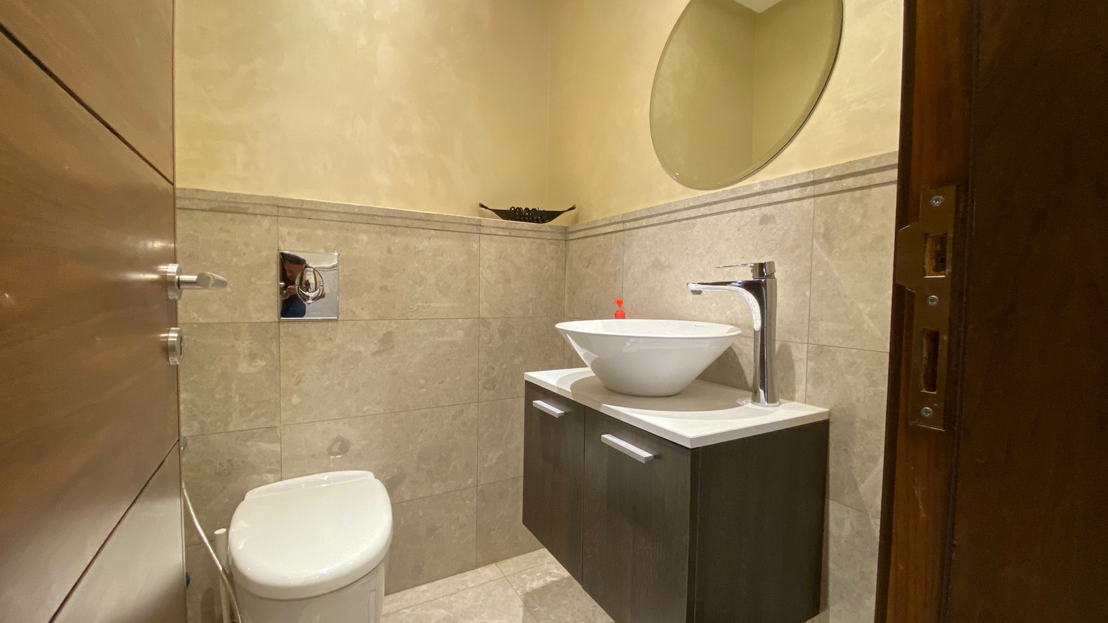 5c8bca00-0848-499b-9cff-7c22a8f92cd5 Contemporary bathroom with a vessel sink on a dark vanity, tall chrome faucet, round mirror, and beige-tile walls and floor.