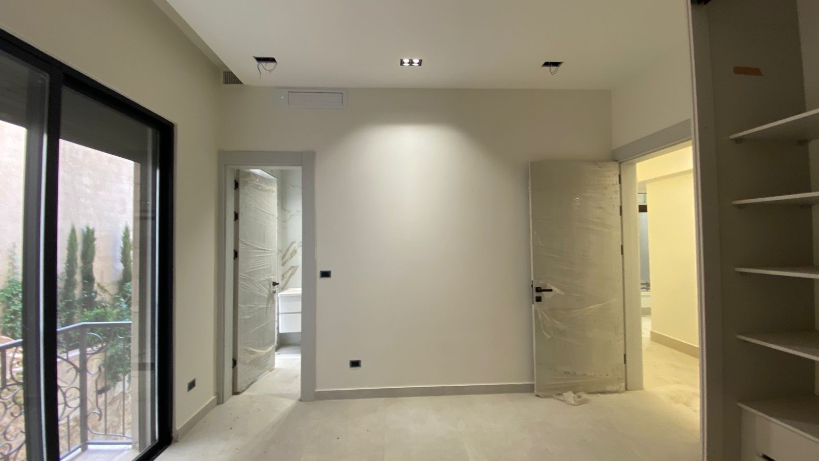 5d8d72a1-7106-4ac0-8621-0f89f667e62b Interior room under renovation with plastic-wrapped doors, a glass balcony door on the left, and built-in shelves on the right, bright white walls.