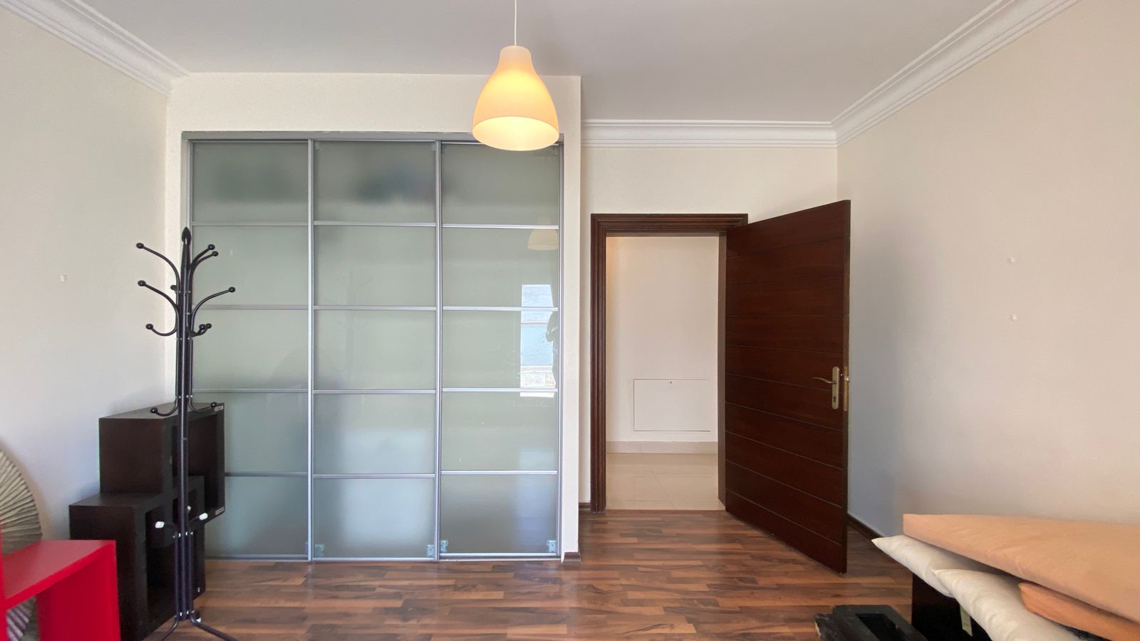 5dc687c5-c8bd-4eb9-9813-ab4fbba33ce4 Living room with frosted glass sliding wardrobe, a black coat rack, a small black shelf, and a red side table near the left wall, wood floor, and an open wooden door to another room.