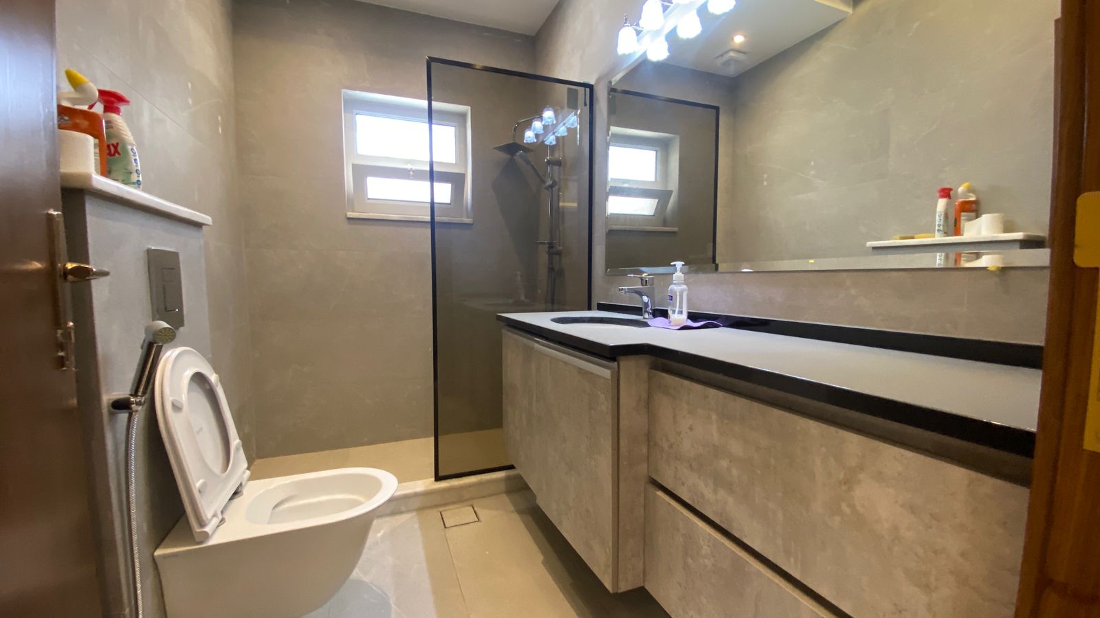 5df84deb-6524-40b7-b846-7405a8267d2a Modern gray bathroom with a toilet, handheld bidet sprayer, glass shower enclosure, and a marble-like vanity with sink and large mirror.