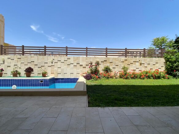 Backyard scene with a blue-tiled rectangular pool, beige stone wall with potted plants, and a wooden fence surrounding a flower bed on the right.