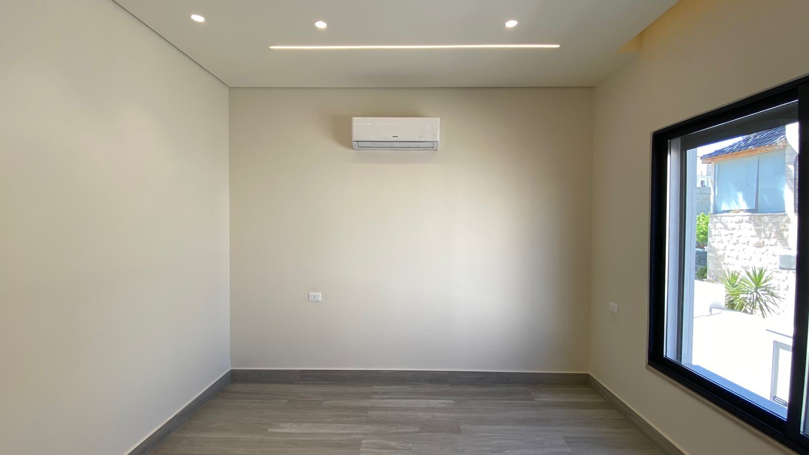 5e57afb9-4bd5-4ab0-9b8e-896093e072ae Empty modern room with light beige walls, gray wood floor, wall air conditioner, and a large right-side window.