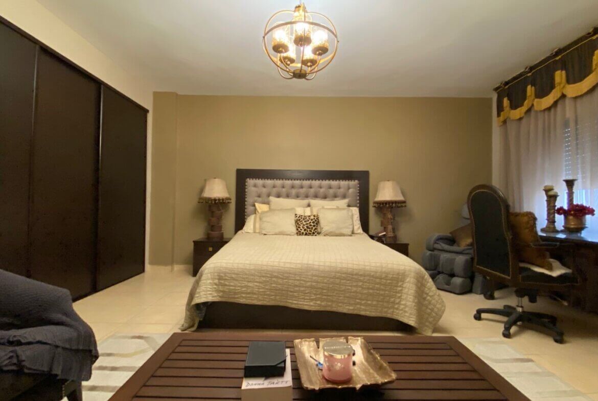 5eacfb12-7ff6-49f2-946a-09bd10a2b89a Bedroom with a king-size bed and tufted headboard, flanked by nightstands and lamps, beside a large dark wood wardrobe on the left.