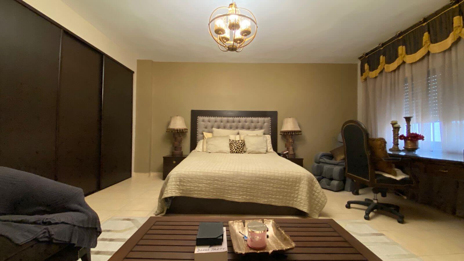 5eacfb12-7ff6-49f2-946a-09bd10a2b89a Bedroom with a king-size bed and tufted headboard, flanked by nightstands and lamps, beside a large dark wood wardrobe on the left.