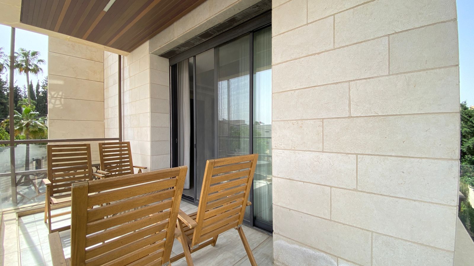 5ed73de2-7871-44e6-af33-4f661416eb3f Outdoor balcony with wooden chairs and a glass railing, beige stone walls, and sliding glass doors. Palm trees visible in the background.