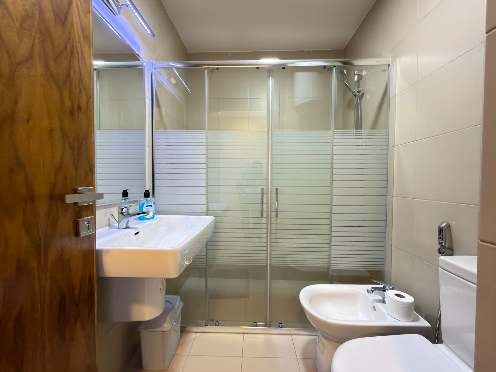 5f53809c-7684-4e2f-8c61-e6f8a67223c4 Shower enclosure with frosted horizontal stripes, beside a white sink and a wooden door; toilet and bidet on the right.