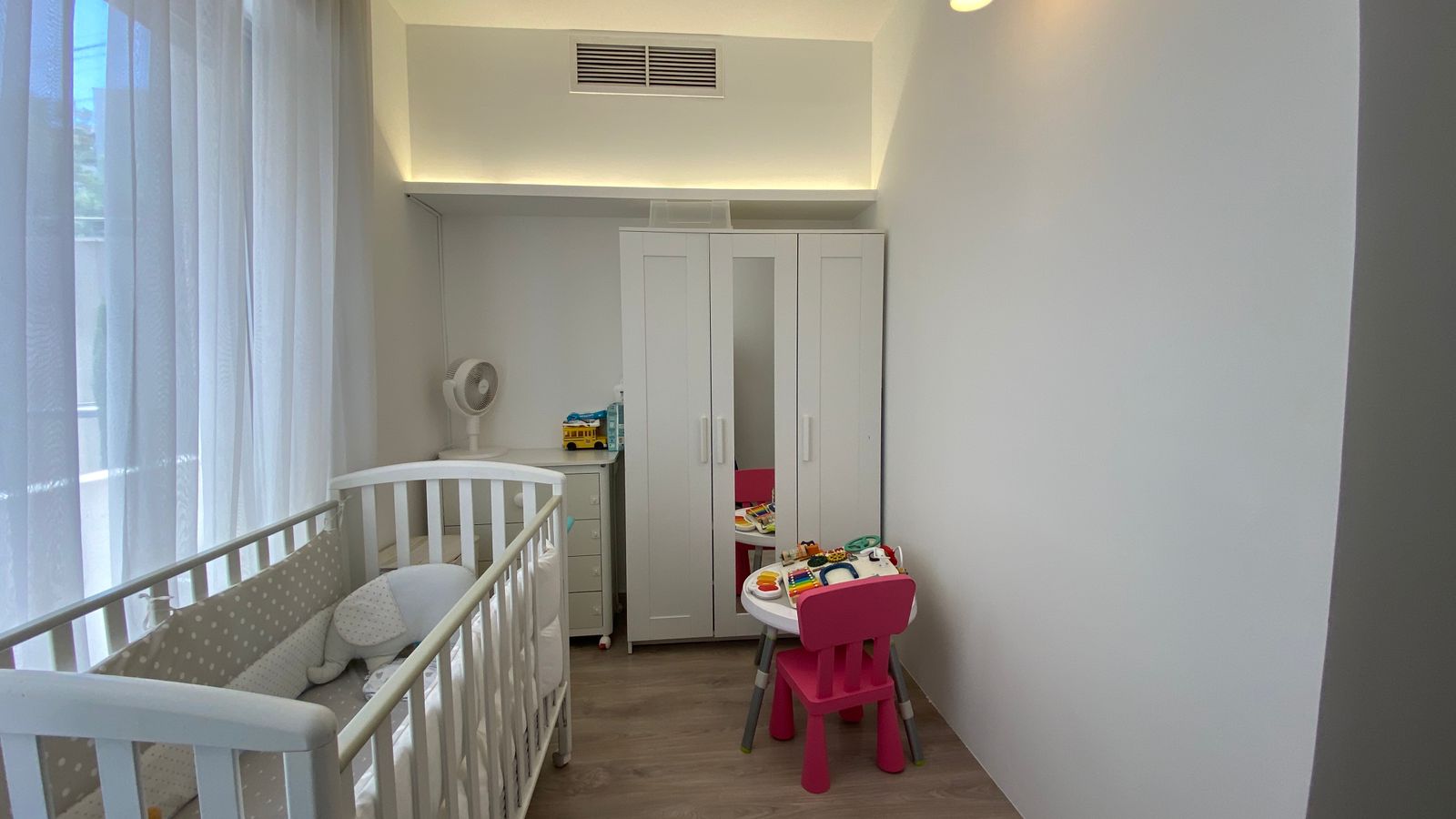 5f627e7b-1b2f-4a50-aaf0-bafc903f6707 Bright white nursery with a crib on the left, sheer curtains, a small dresser with a fan, and a tall white wardrobe with a mirrored door; pink table and chairs with toys nearby.