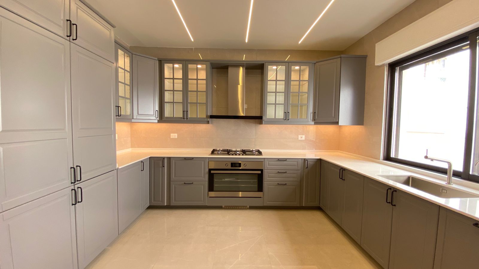 5f7fdd96-d703-4db1-ae2a-77493c43ae13 L-shaped modern kitchen with gray cabinets, glass-front upper cabinets, stainless steel oven and gas cooktop, and a window over the sink with a range hood above the stove.