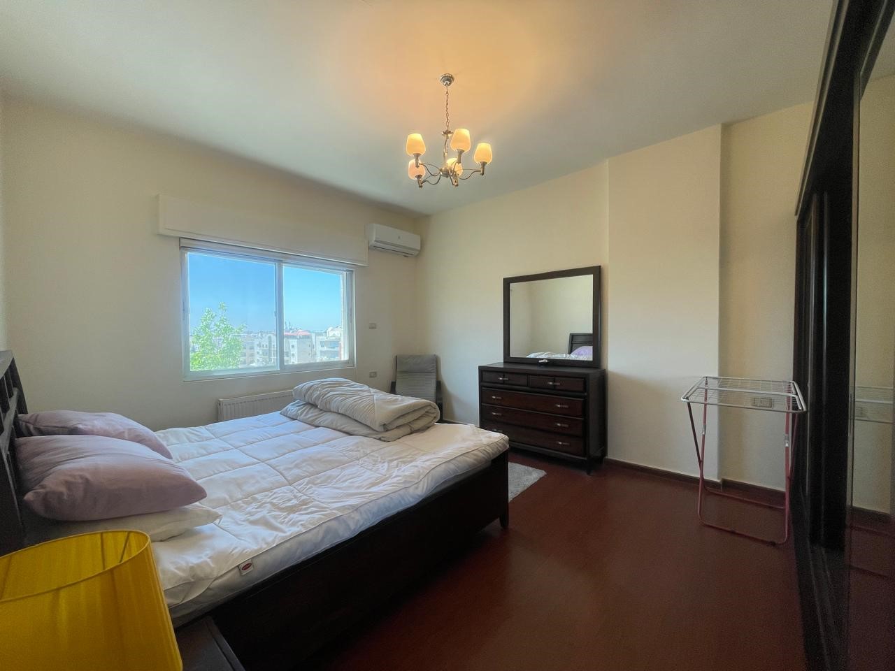5faa3912-d952-4e17-a741-7b27839d9d87 Dark wooden bed with white quilt and pink pillows, facing a dresser with a mirror in a bright bedroom