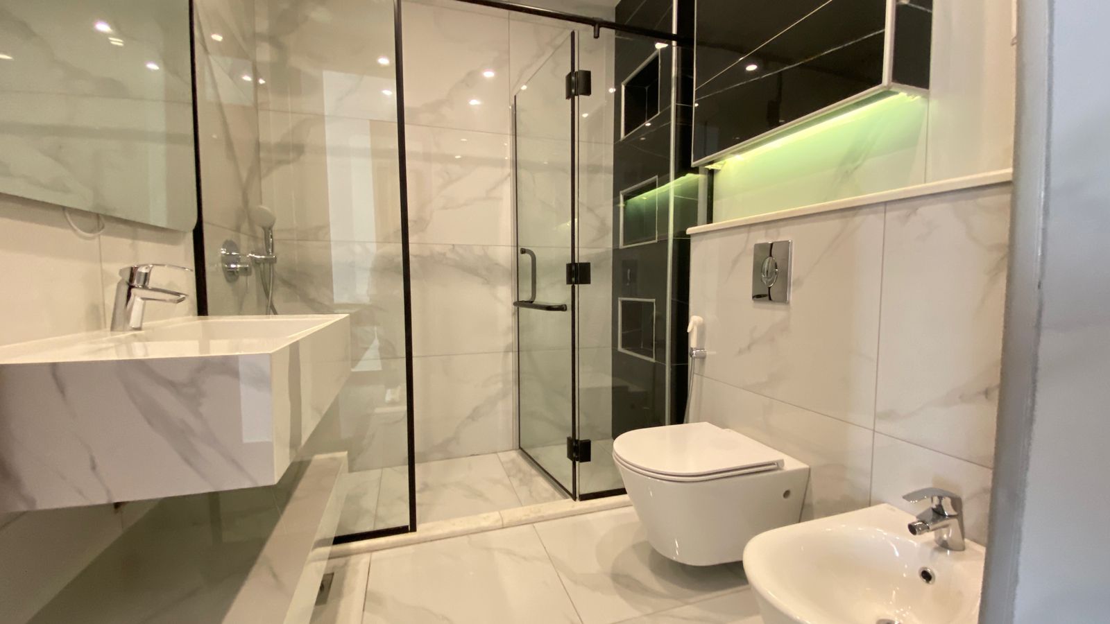 5fdbb9aa-41ba-4813-b89c-927eaa35c81f Modern white marble bathroom with glass shower enclosure, wall-mounted sink, wall‑hung toilet, and bidet with soft green lighting behind mirrored cabinet.