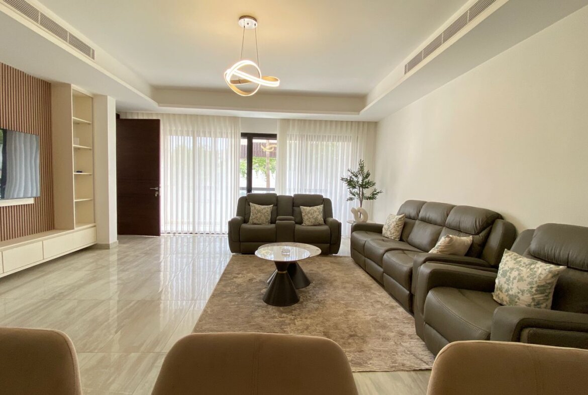 Spacious living room with gray leather sectional, matching loveseat, round coffee table, and a rug near glass doors with sheer curtains.
