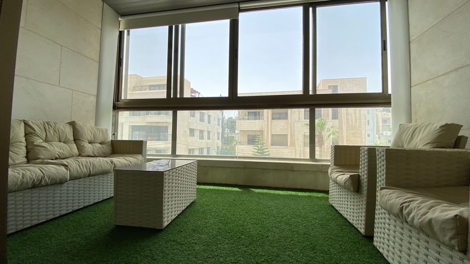 602a82e9-9671-4321-9b98-3c22132482e7 Enclosed balcony lounge with white wicker sofa and armchairs, beige cushions, a glass coffee table, and a green artificial grass floor.