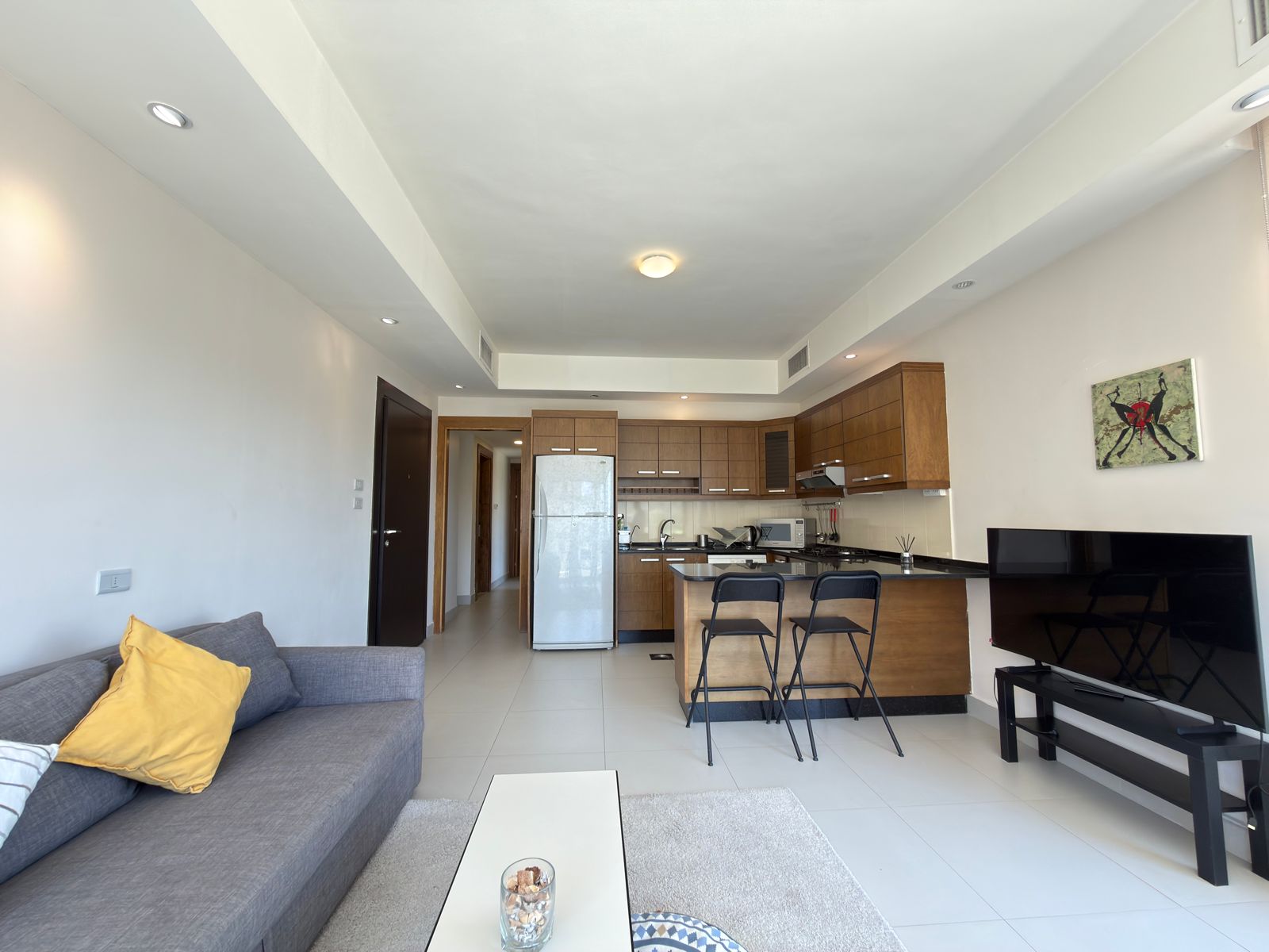 60486cea-ca5b-49f1-850f-bed9164b64ca Open-plan living area with a gray sofa and yellow cushions, adjacent kitchen with wooden cabinets and a stainless-steel fridge. A black TV sits on a stand to the right.