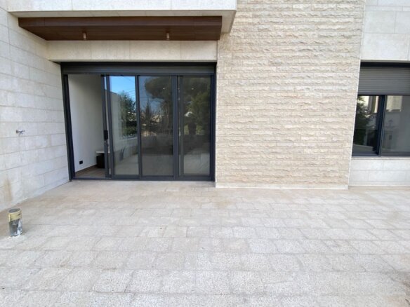 Wide tiled terrace in front of a beige stone building, featuring large sliding glass doors and a window with a rolling shutter on the right.