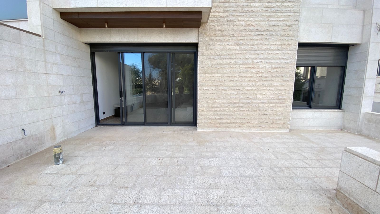 6097d466-4d32-463e-b063-fc026ad12545 Wide tiled terrace in front of a beige stone building, featuring large sliding glass doors and a window with a rolling shutter on the right.