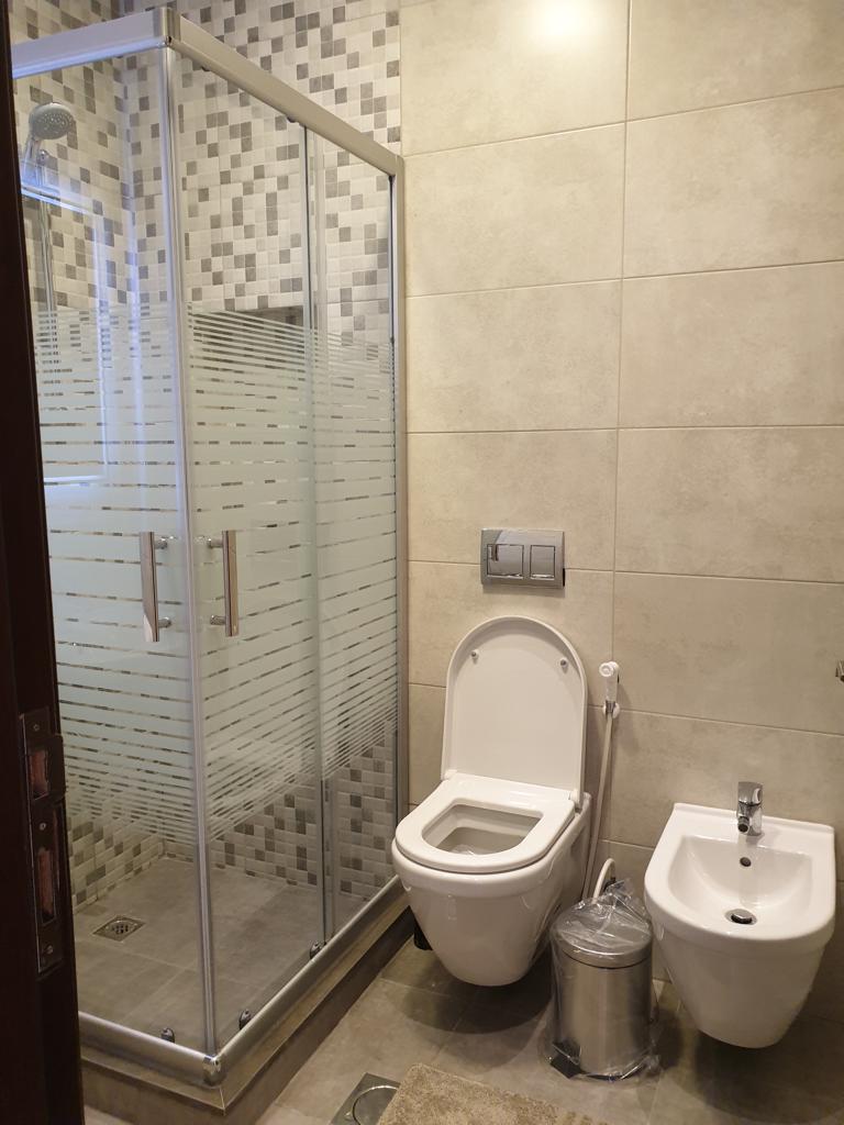 6130d15a-8ac9-41a7-af36-190edc7aa36a Modern bathroom with a glass-enclosed shower, wall tiles, a wall-mounted dual-flush toilet, a nearby bidet, a stainless trash can, and a beige bath mat.
