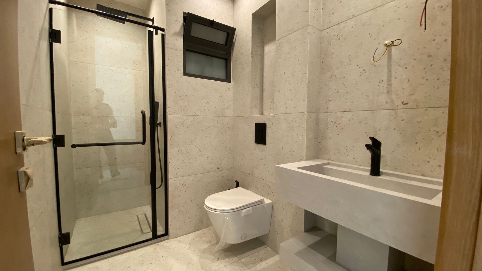 61720329-2d1d-4703-a25b-3280efb9fbc4 Modern bathroom with glass-framed shower, wall-mounted toilet, and concrete-like tiles; exposed wires near the sink suggest ongoing construction/finish work.