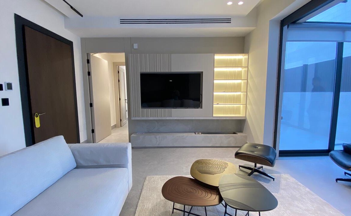 Modern living room with a wall-mounted TV, backlit shelves, and a built-in TV console; neutral sofa and sculpted coffee tables.