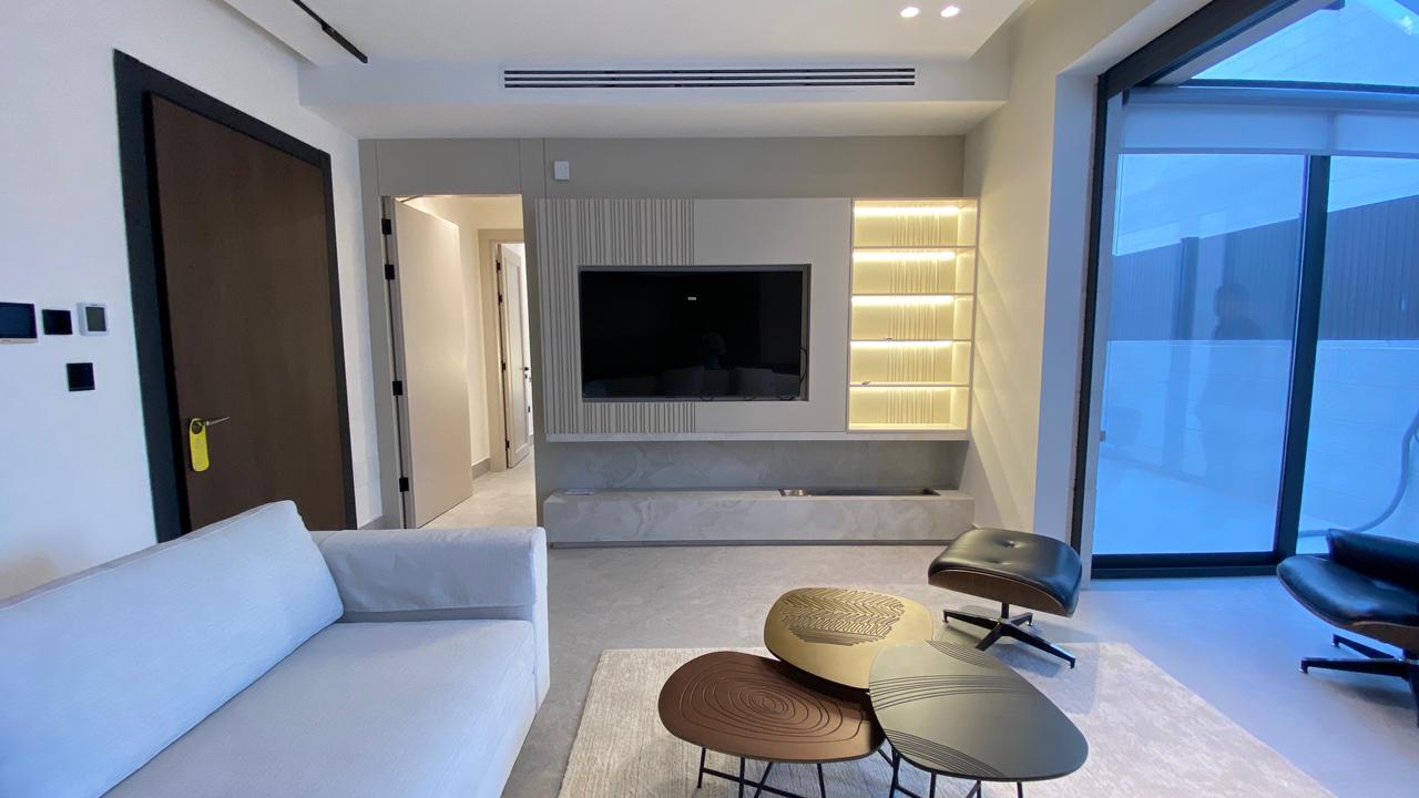 Modern living room with a wall-mounted TV, backlit shelves, and a built-in TV console; neutral sofa and sculpted coffee tables.