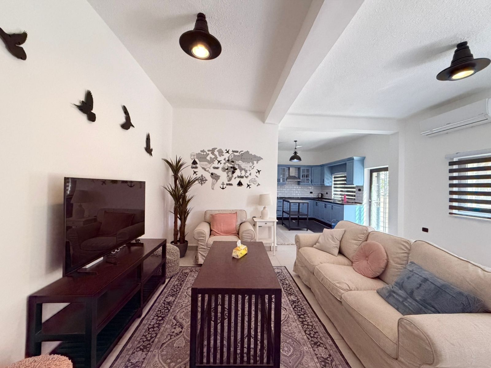 6244a06e-6e09-44d4-b218-2f30776f72f1 Living room with beige sofa and coffee table, TV on a dark stand, and a wall map with bird silhouettes; blue kitchen in the background.
