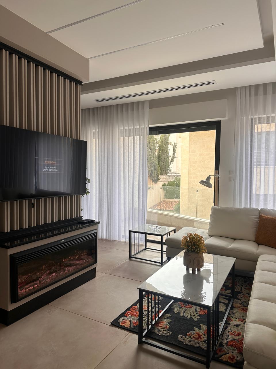 62922a13-53d1-4108-a5f6-37dac2a4a595 Living room with a wall-mounted TV above a linear fireplace, beige sectional sofa, and two glass-top coffee tables near a floral rug.