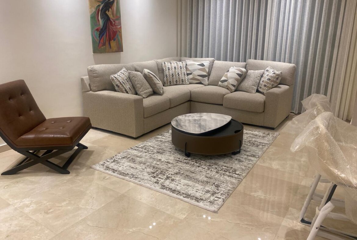 L-shaped beige sectional with patterned cushions, round marble-top coffee table, brown leather lounge chair, and sheer curtains in a modern living room.